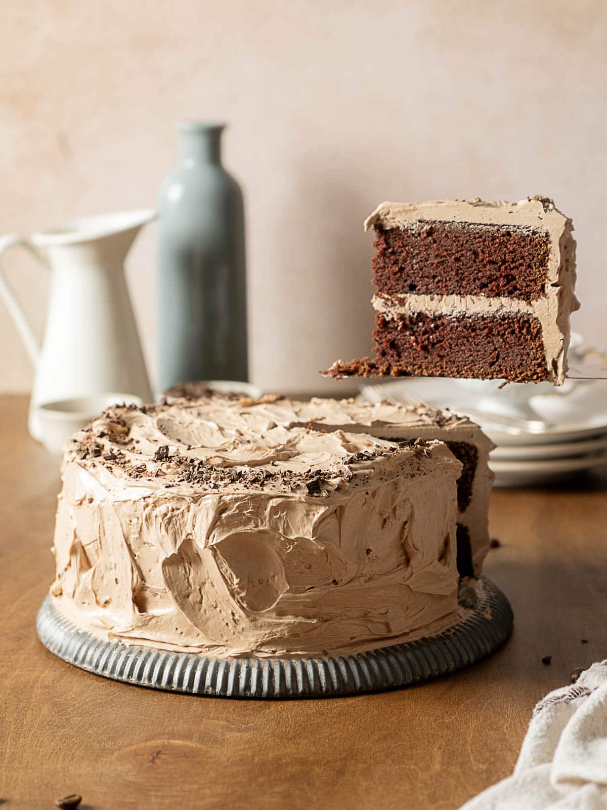 A chocolate layer cake with chocolate frosting sits on a metal platter. A slice is being lifted, showing the cake’s interior. Plates, a vase, and a pitcher are in the background.
