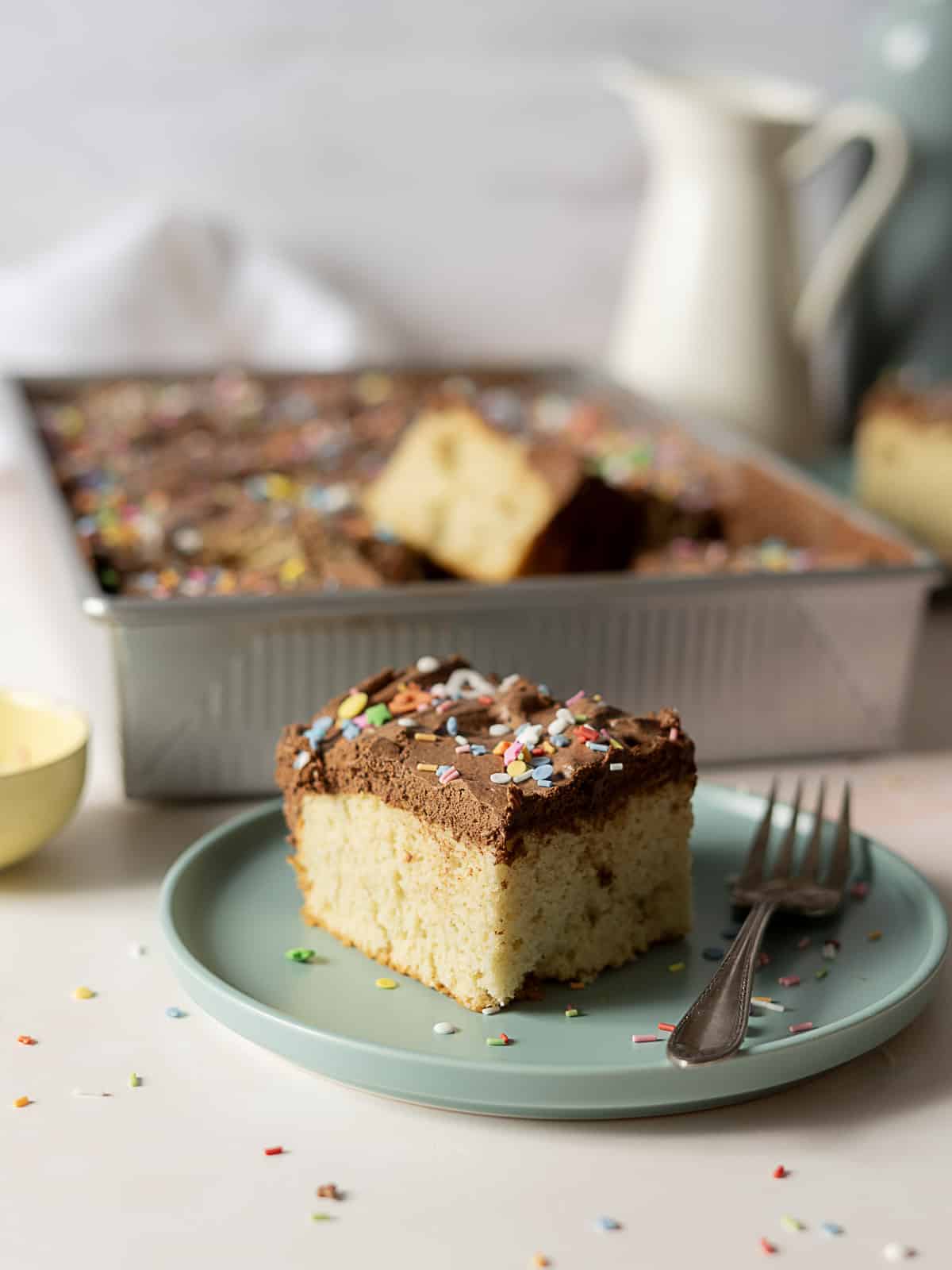 A slice of yellow cake with chocolate frosting and colorful sprinkles sits on a green plate with a fork, with the rest of the cake in a pan in the background.
