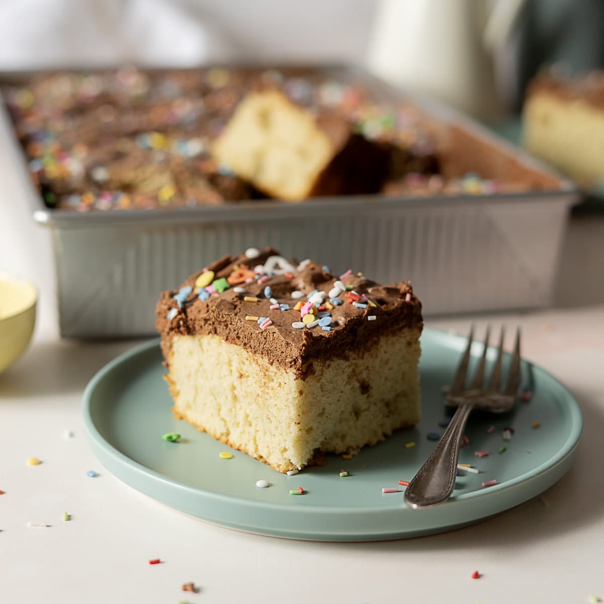 A slice of yellow cake with chocolate frosting and sprinkles sits on a blue plate with a fork, with the rest of the cake in a pan in the background.