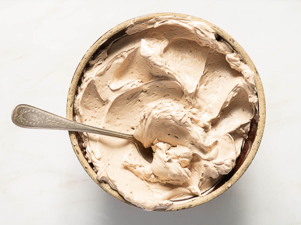 A bowl of whipped light brown frosting with a spoon in it, viewed from above on a light surface.