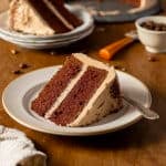 A slice of chocolate layer cake with light brown frosting on a white plate with a fork, sitting on a wooden table.