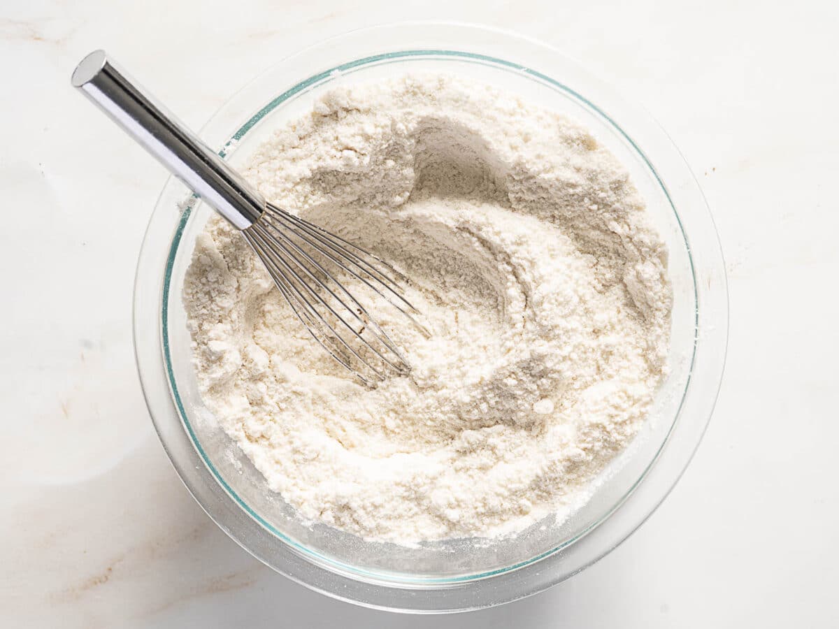 A metal whisk rests in a glass bowl filled with mixed flour on a white surface.