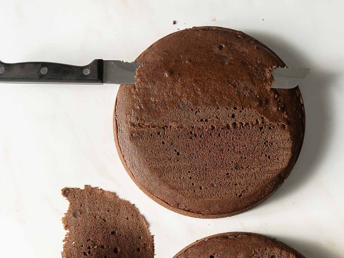 A chocolate cake with the domed top being leveled off using a serrated knife; the removed cake top is on the side.