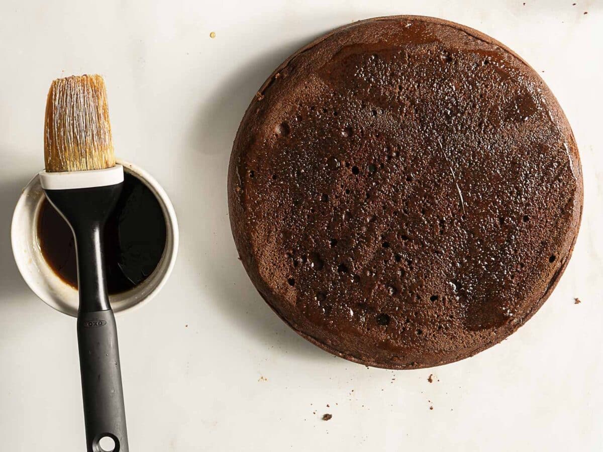 A round chocolate cake with a shiny surface sits next to a brush resting in a small bowl of liquid on a white background.