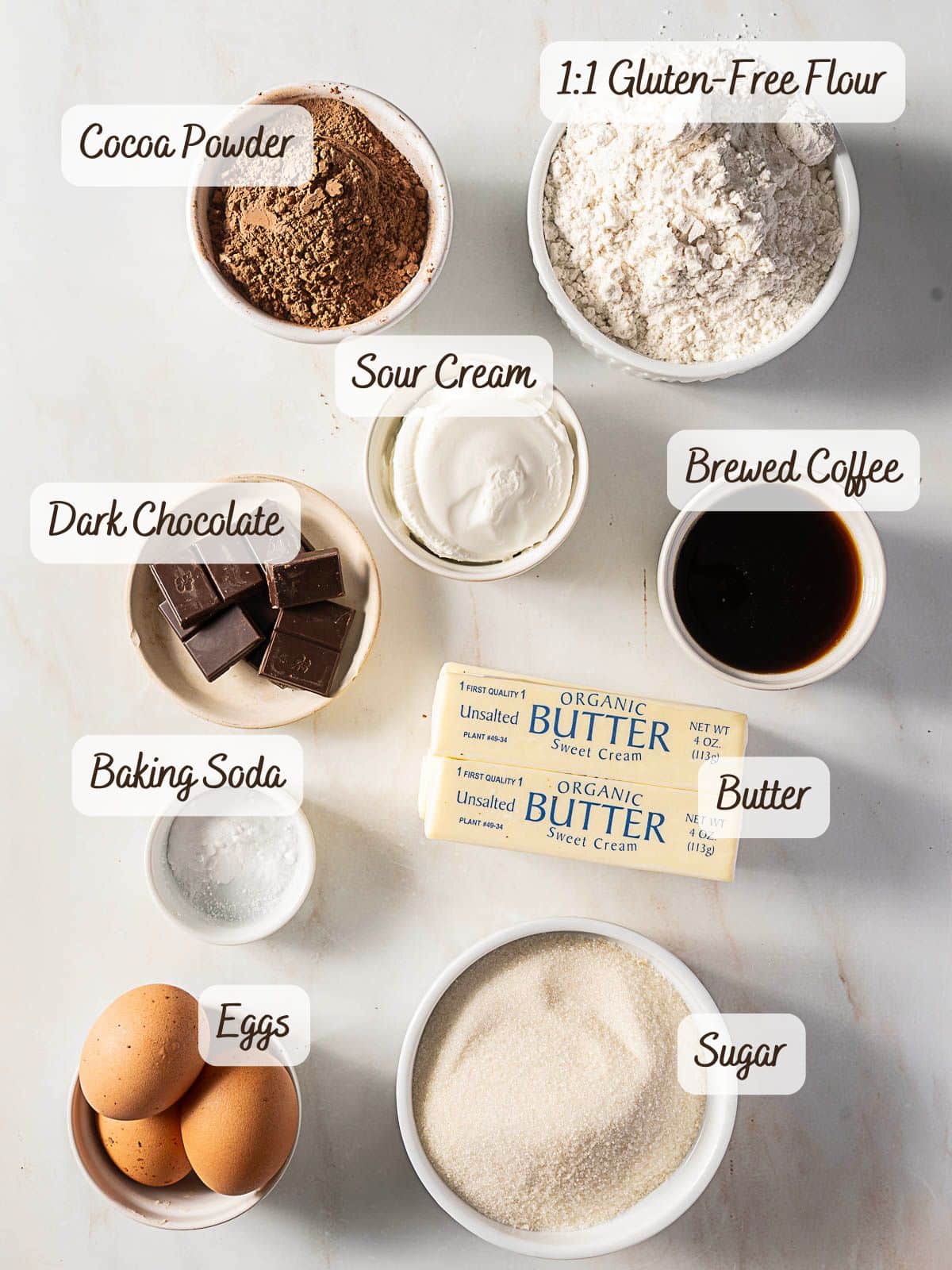 A flat lay of baking ingredients in bowls and plates for a gluten free mocha cake: cocoa powder, gluten-free flour, sour cream, brewed coffee, dark chocolate, butter, baking soda, eggs, and sugar.