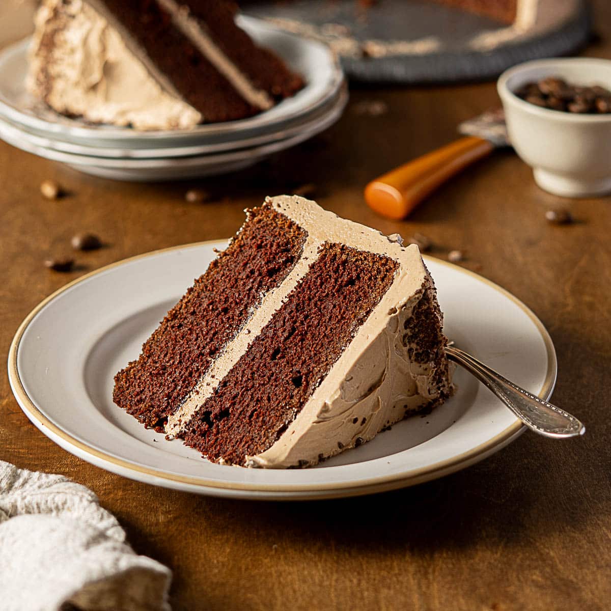 A slice of chocolate layer cake with light brown frosting on a white plate with a fork, sitting on a wooden table.