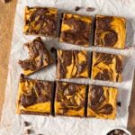 Nine pumpkin swirl brownies are arranged in a grid on parchment paper, with one brownie showing a bite taken out. Chocolate chips are scattered around.