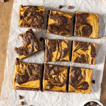 Nine pumpkin swirl brownies are arranged in a grid on parchment paper, with one brownie showing a bite taken out. Chocolate chips are scattered around.