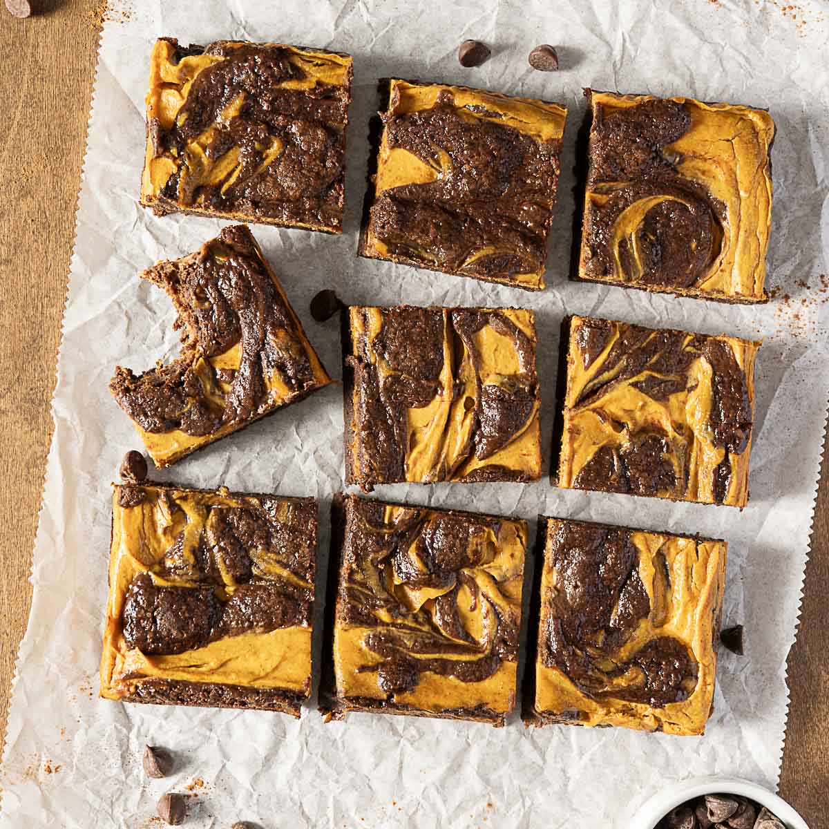Nine pumpkin swirl brownies are arranged in a grid on parchment paper, with one brownie showing a bite taken out. Chocolate chips are scattered around.