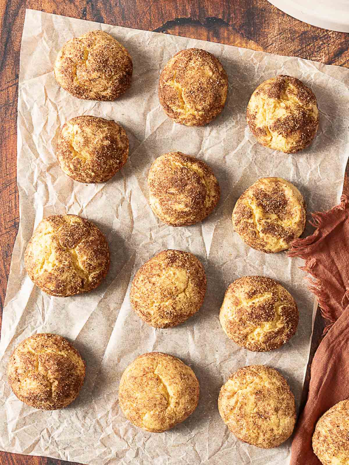 Twelve round snickerdoodle cookies arranged on a sheet of parchment paper on a wooden surface.