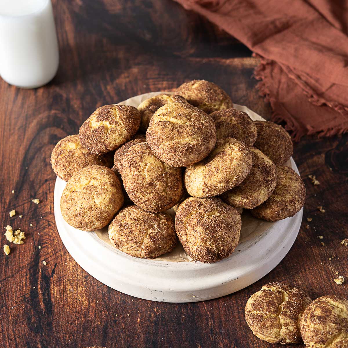 A plate of round snickerdoodle cookies is arranged on a wooden surface, with a glass of milk and a brown cloth in the background.