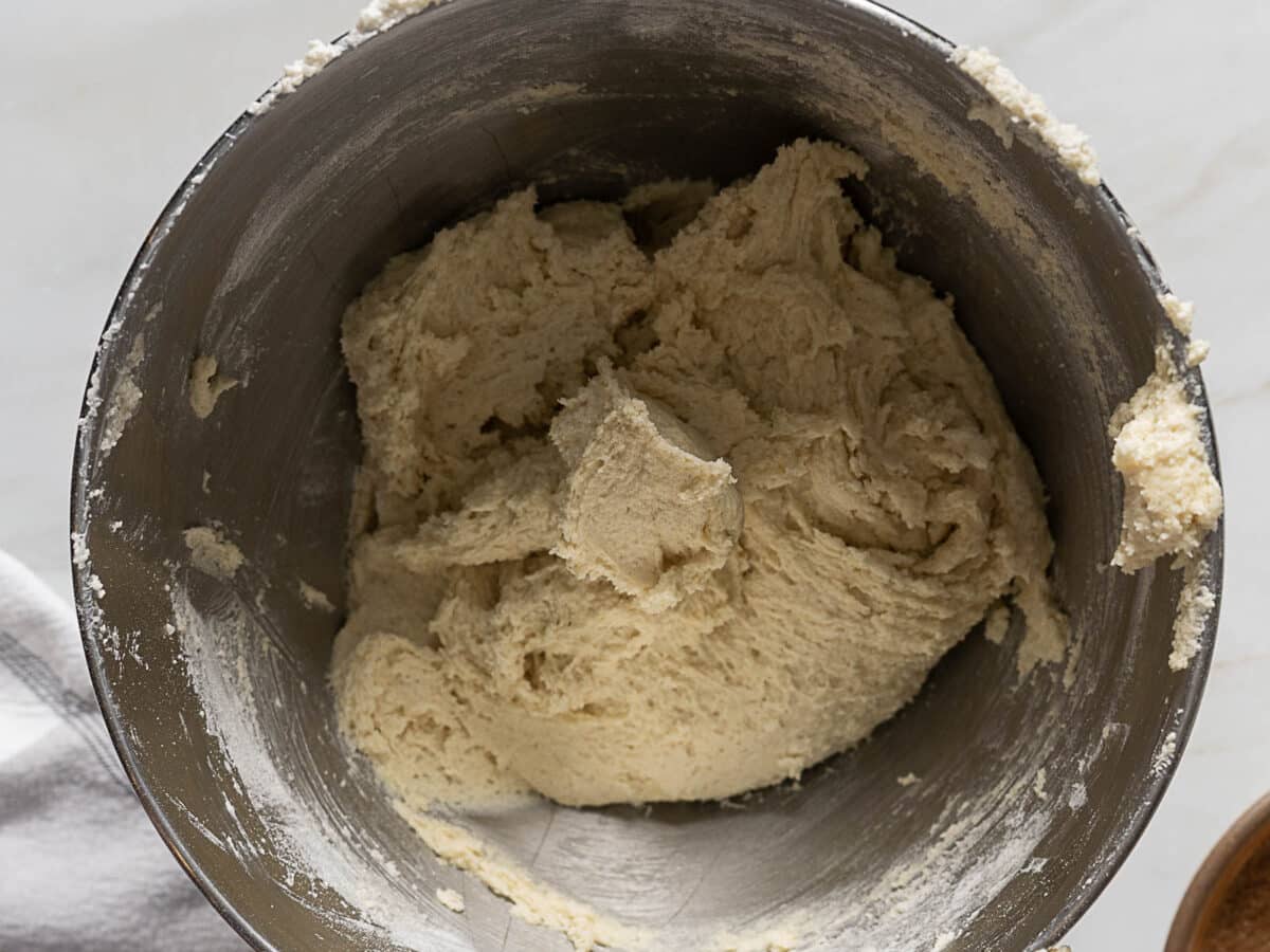 A metal mixing bowl filled with an off-white, rough-textured dough mixture on a light-colored surface.