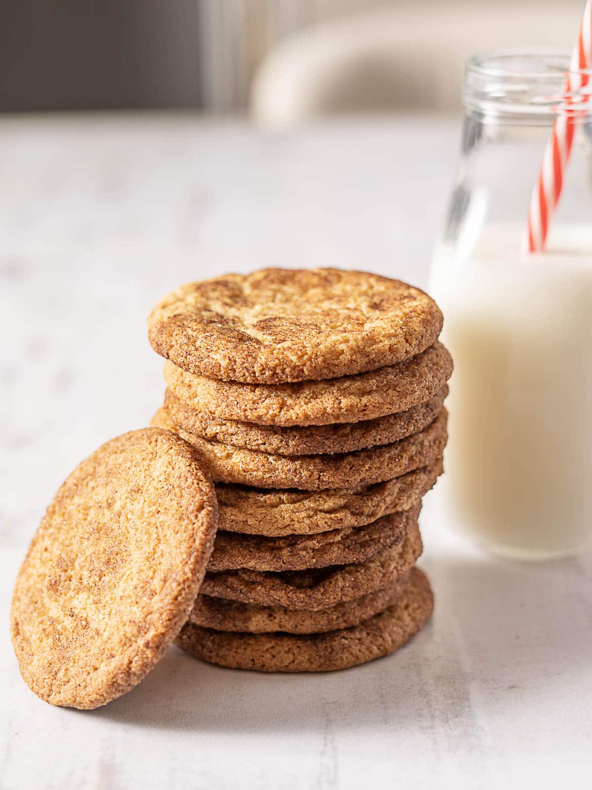 A stack of snickerdoodle cookies sits on a white surface next to a bottle of milk with a red and white striped straw.
