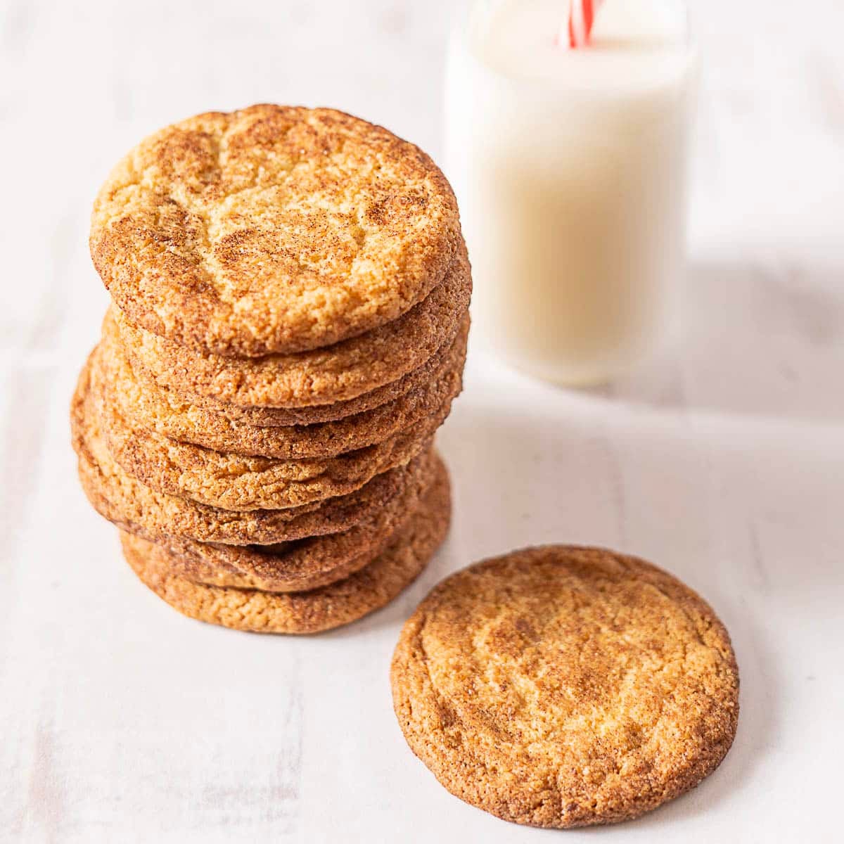 A stack of snickerdoodle cookies sits next to a single cookie and a glass of milk with a red-striped straw on a white surface.