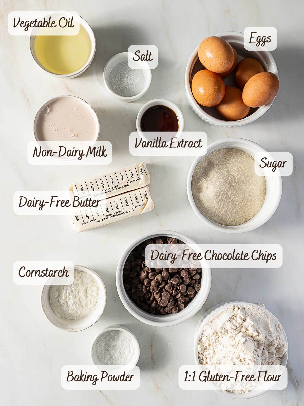 Assorted baking ingredients on a white surface, perfect for making a delicious gluten free yellow cake—includes eggs, sugar, vegetable oil, non-dairy milk, vanilla extract, salt, dairy-free butter, gluten-free flour, baking powder, cornstarch, and chocolate chips.