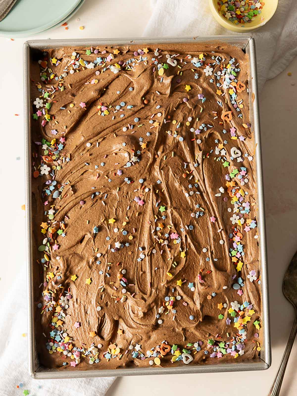 A sheet cake topped with swirled chocolate frosting and colorful sprinkles sits in a metal baking pan on a light surface.