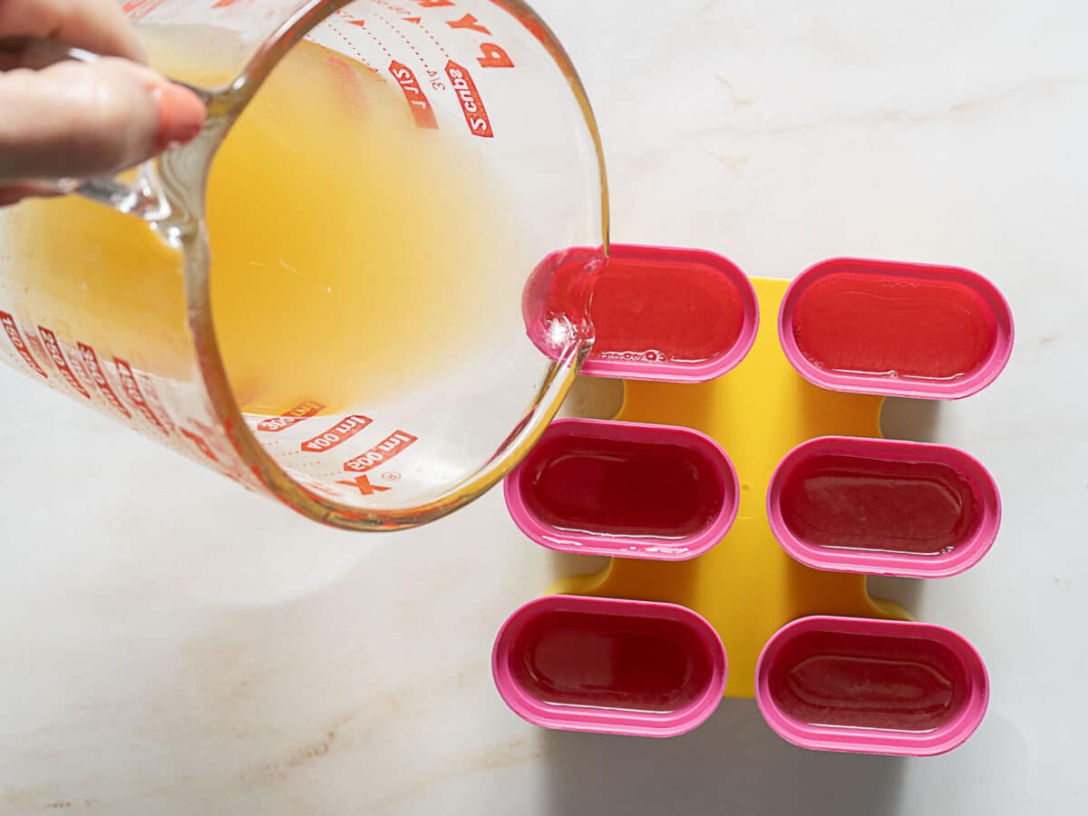 A person pours yellow liquid from a measuring cup into a pink and yellow popsicle mold on a white surface.