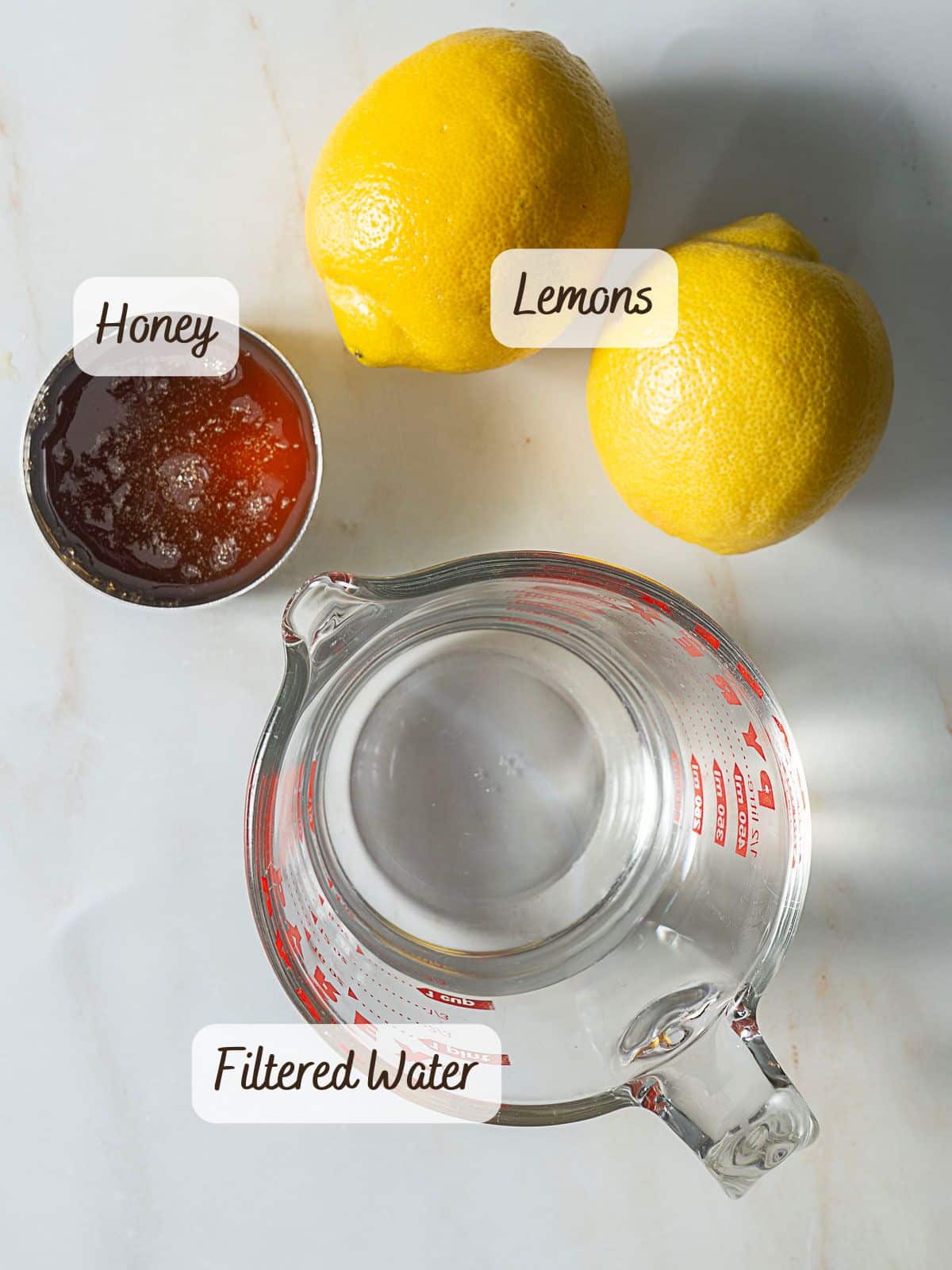 A measuring cup of filtered water, two whole lemons, and a small bowl of honey are arranged on a white surface, each labeled with text—perfect ingredients for refreshing homemade lemon popsicles.