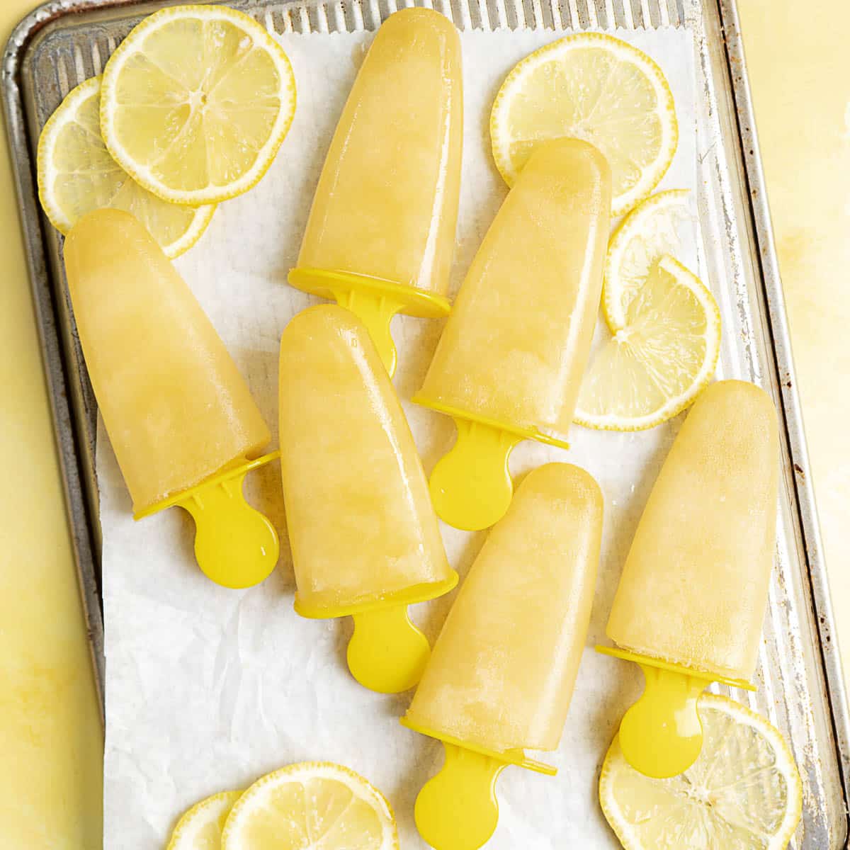 Six yellow popsicles arranged on a parchment-lined baking sheet with several lemon slices scattered around.