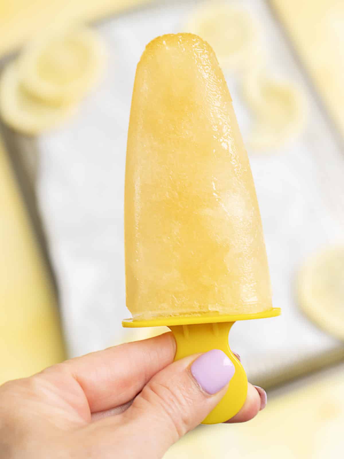 A hand with lavender nail polish holds a yellow popsicle, with blurred lemon slices and a tray in the background.