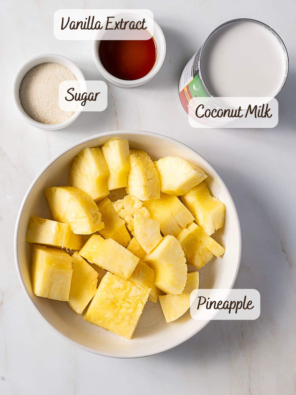A bowl of pineapple chunks, a small bowl of sugar, vanilla extract, and an open can of coconut milk on a white surface—everything you need to make delicious pina colada popsicles, each ingredient clearly labeled.