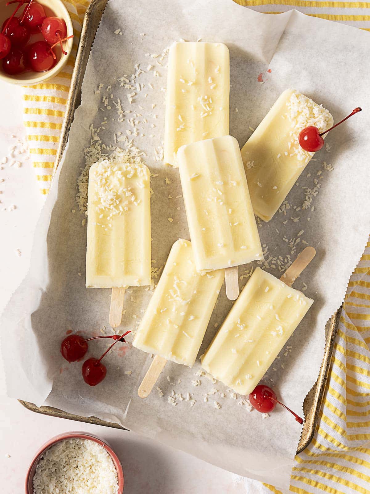 Six creamy popsicles topped with shredded coconut are arranged on parchment paper in a tray, with maraschino cherries scattered around and a bowl of shredded coconut nearby.