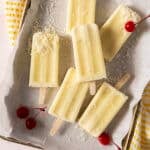 Six creamy yellow popsicles with shredded coconut topping are arranged on parchment paper, with three maraschino cherries nearby.
