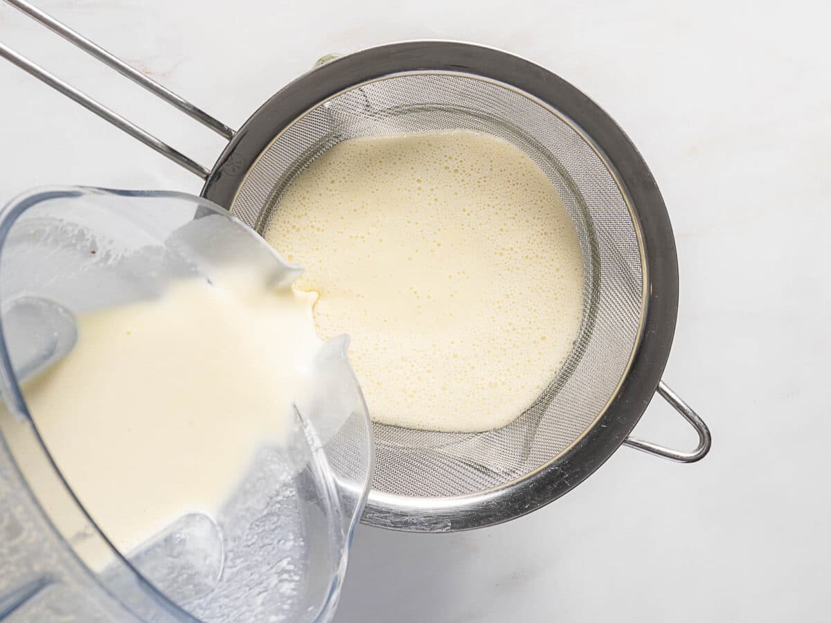 Blended liquid is being poured from a blender through a fine mesh strainer into a bowl on a white surface.