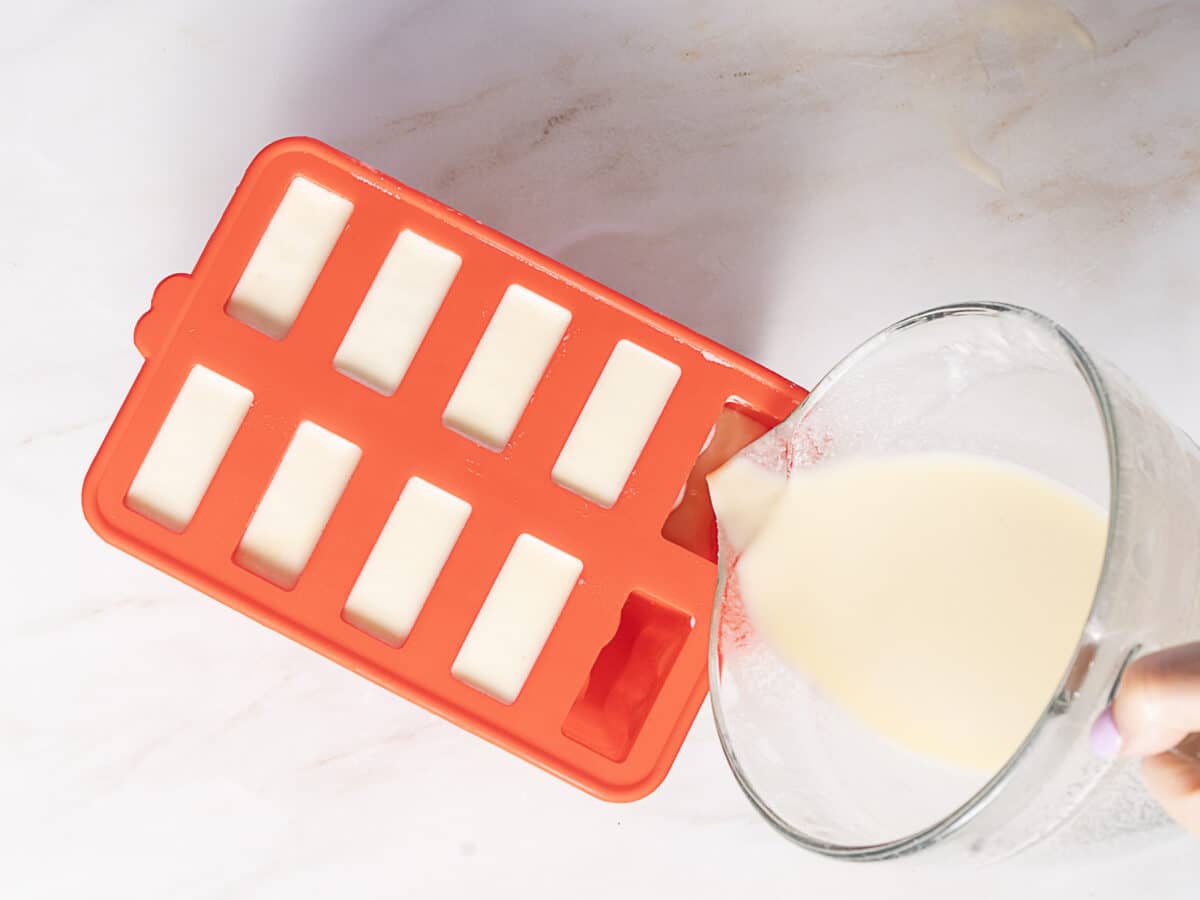 A person pours a creamy liquid from a glass bowl into a red silicone rectangular mold on a white surface.