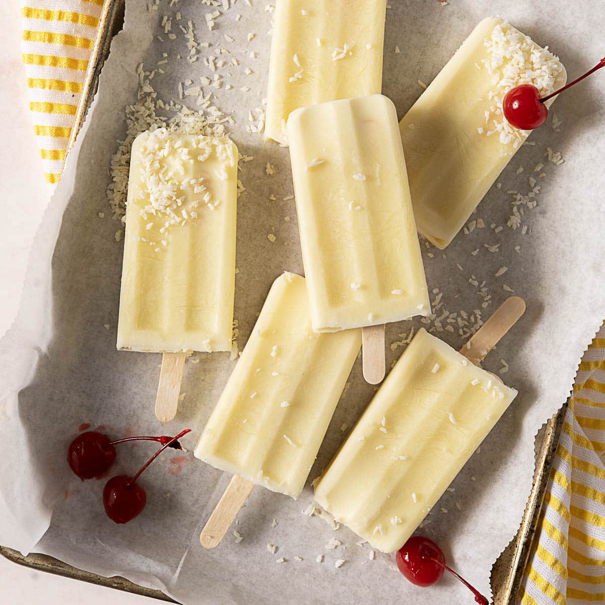 Six creamy yellow popsicles with shredded coconut topping are arranged on parchment paper, with three maraschino cherries nearby.