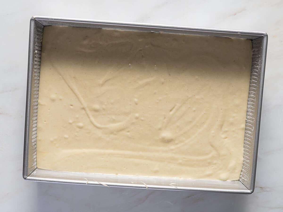 A rectangular metal baking pan filled with an even layer of light-colored cake batter sits on a white surface.