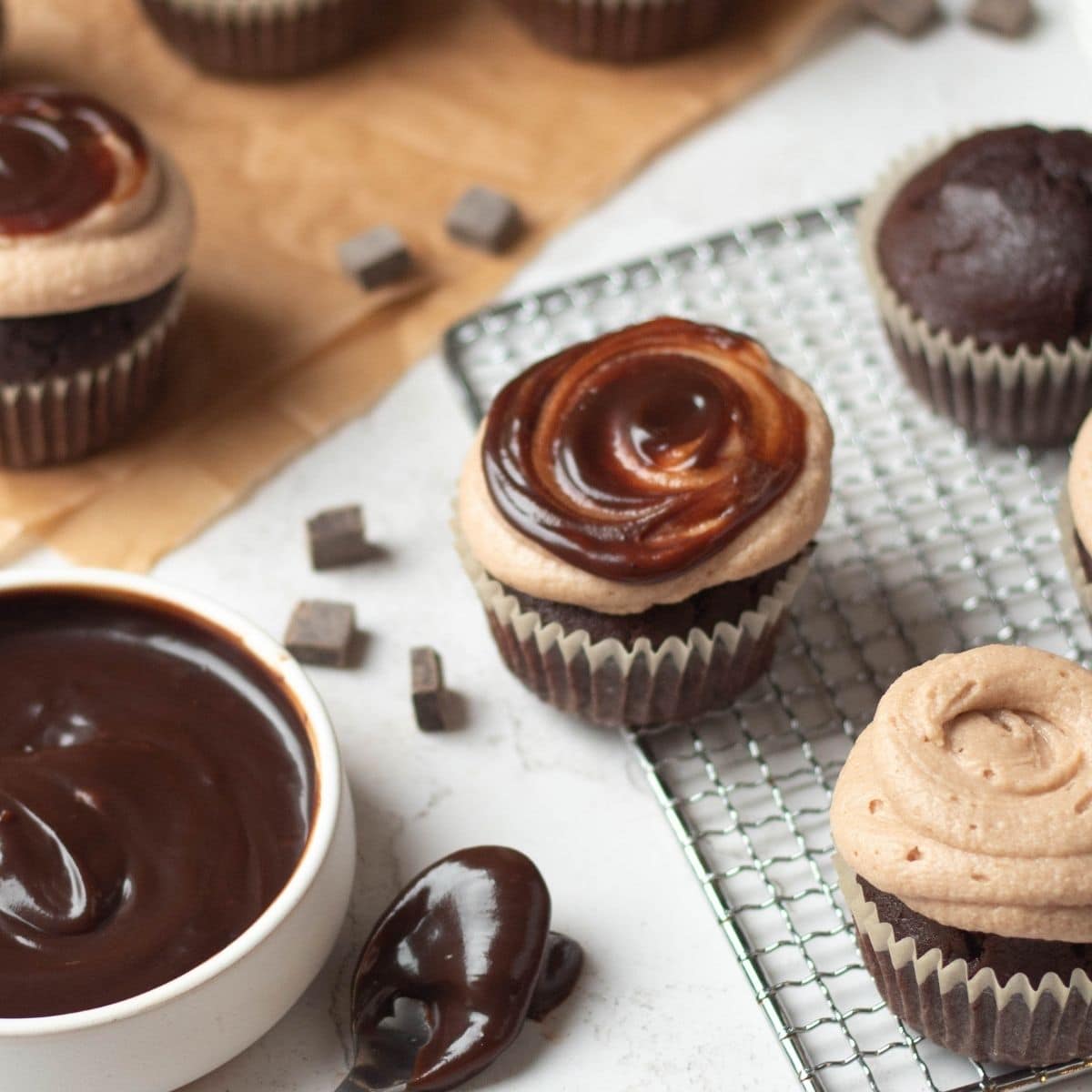 Chocolate cupcakes with two types of frosting, some topped with chocolate ganache and others with chocolate buttercream, are placed on a cooling rack and a white surface.