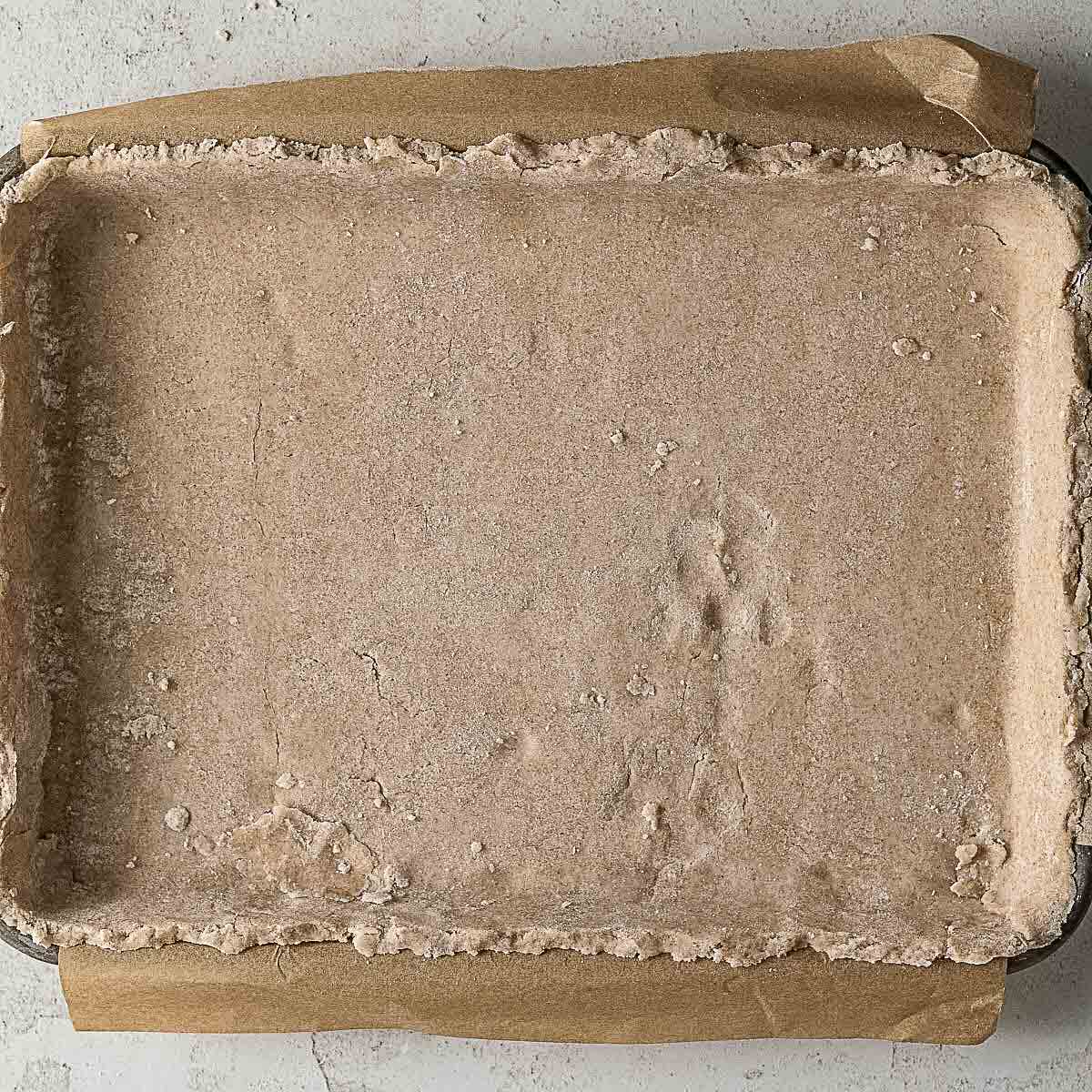 A baking tray lined with parchment paper and filled with an evenly pressed, uncooked dough covering the bottom and sides.