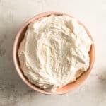 A bowl filled with fluffy, white buttercream frosting sits on a light-colored surface.