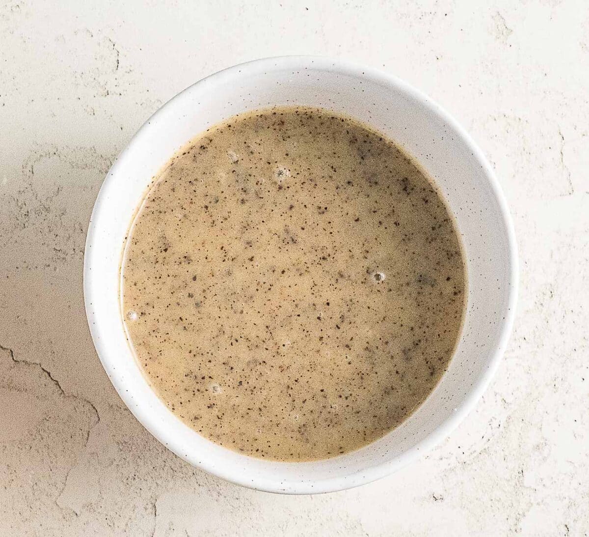 A white bowl filled with a light brown, speckled liquid mixture sits on a textured off-white surface.