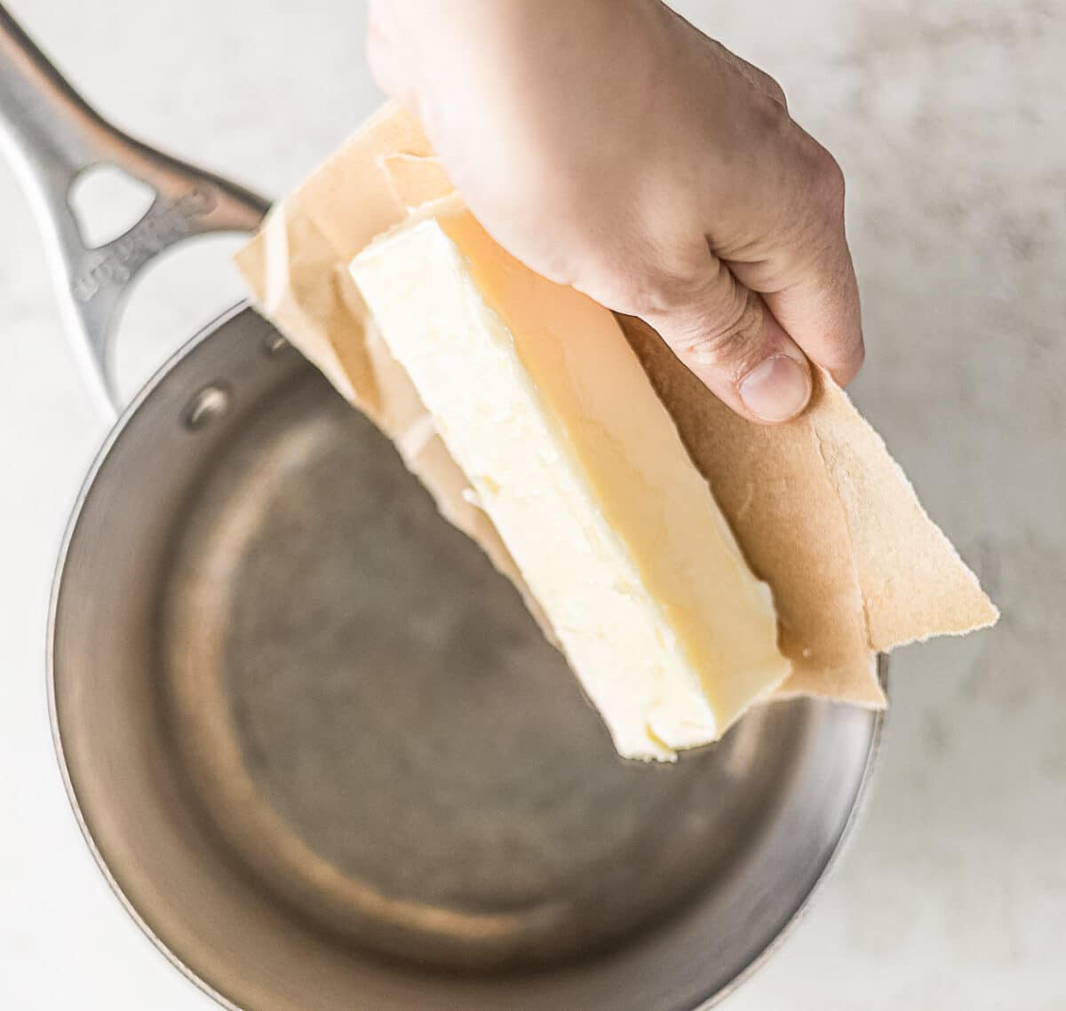 A hand holds a stick of butter above an empty saucepan, partially wrapped in paper.