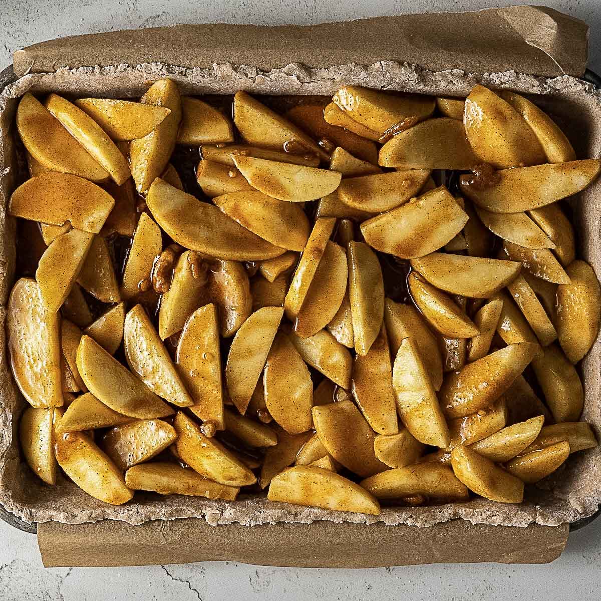 A baking tray lined with parchment paper, filled with sliced apples coated in cinnamon and sugar, ready to be baked.