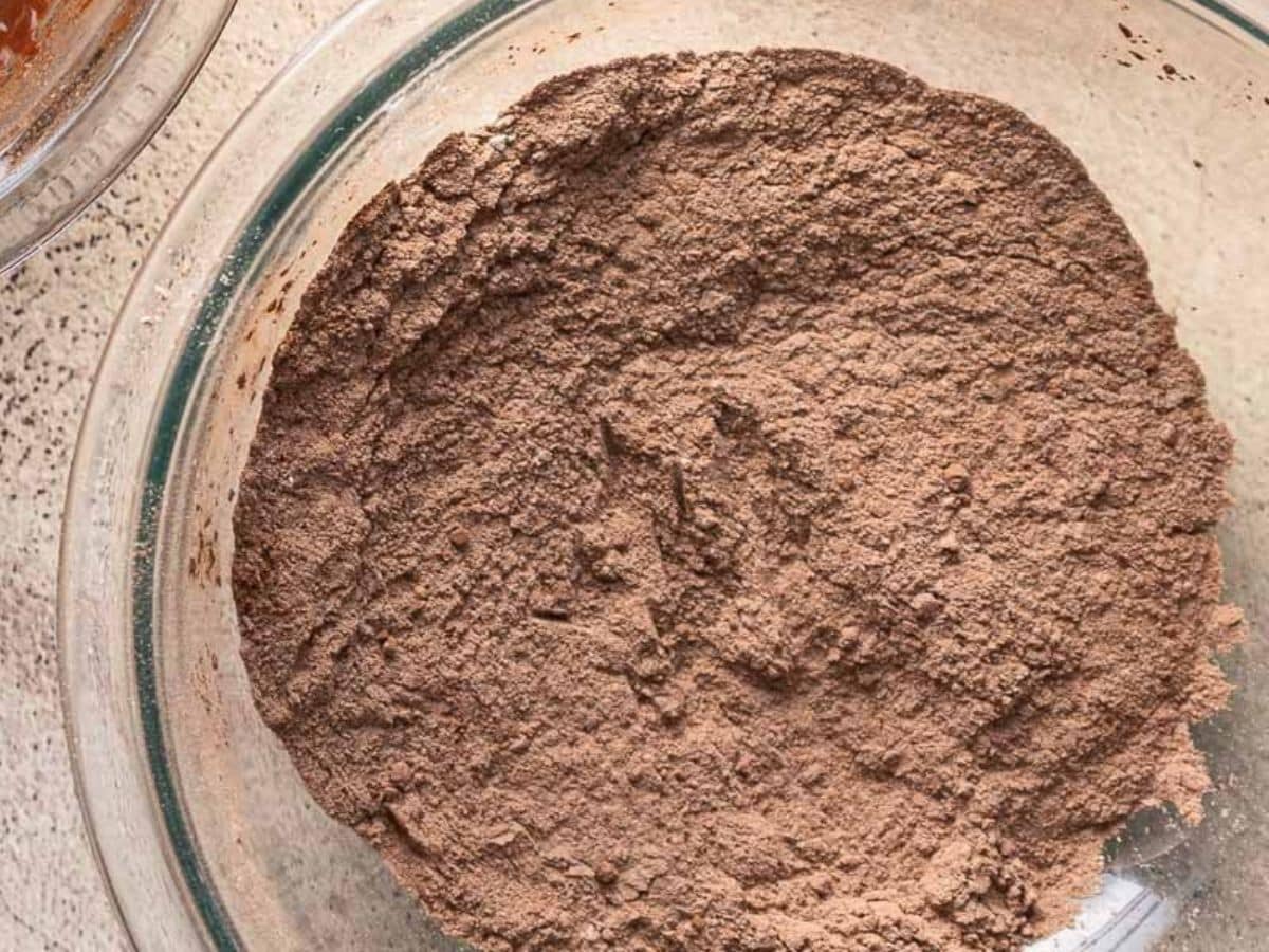 A glass bowl filled with a mixture of dry chocolate powder ingredients, viewed from above on a light surface.