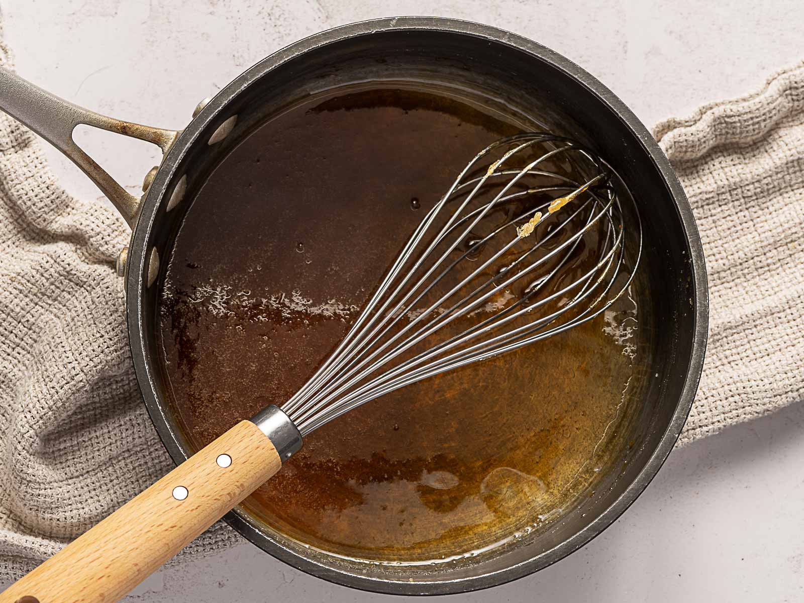 A saucepan with amber melted sugar and a metal whisk resting inside, placed on a beige textured cloth on a light surface.