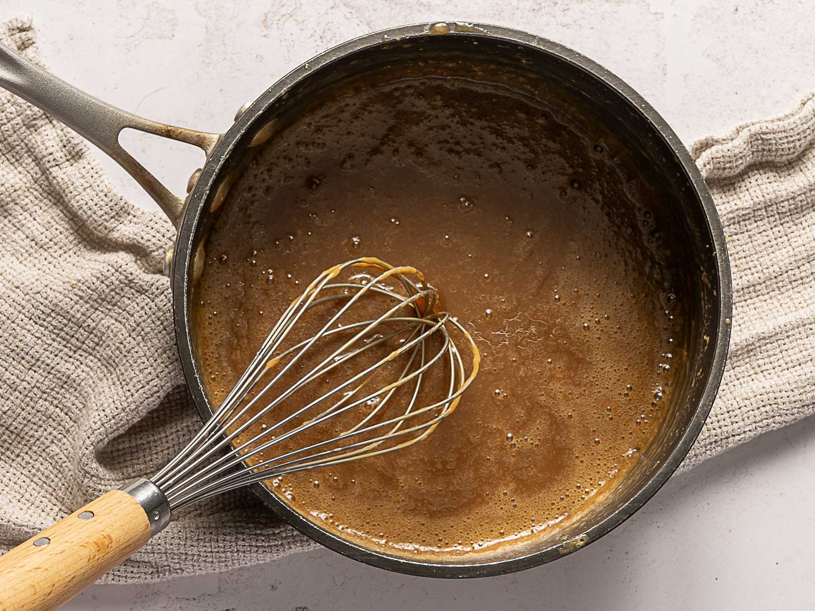 A saucepan of rich salted caramel sauce being whisked sits on a textured beige cloth.