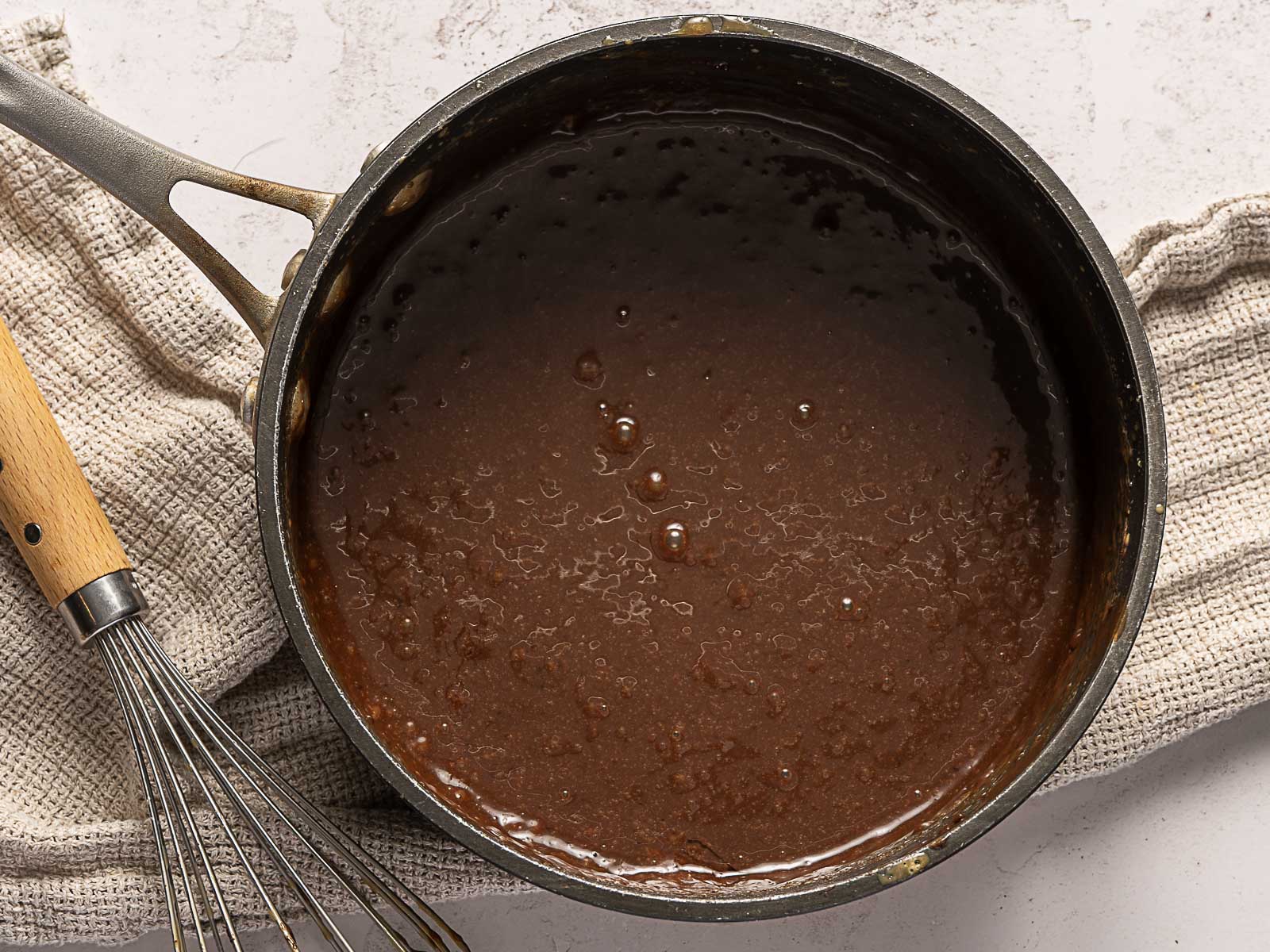 A saucepan filled with a dark, glossy chocolate caramel sauce sits on a textured towel beside a metal whisk on a light-colored surface.