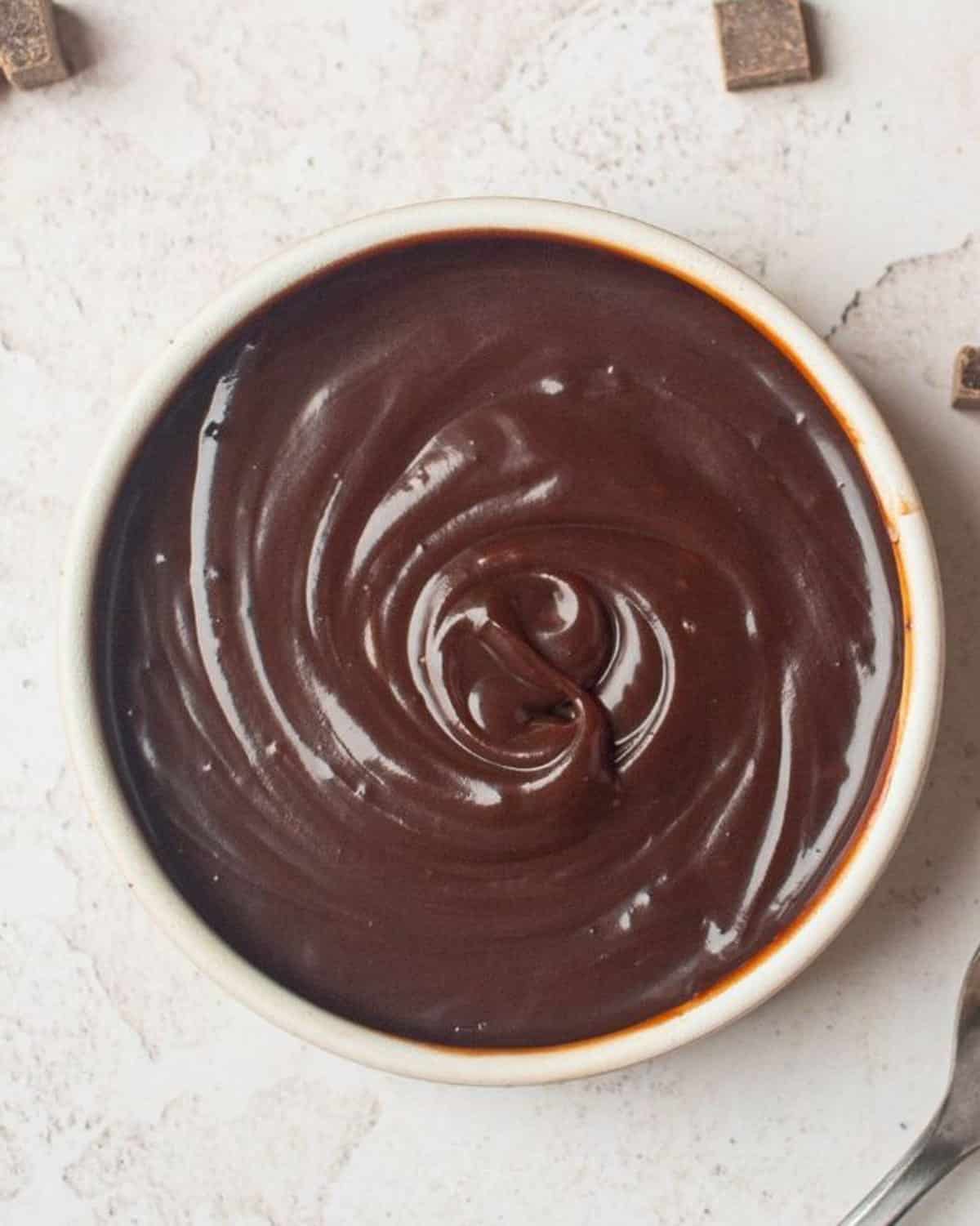 A bowl filled with smooth, swirled chocolate ganache, viewed from above on a light surface with chocolate pieces nearby.