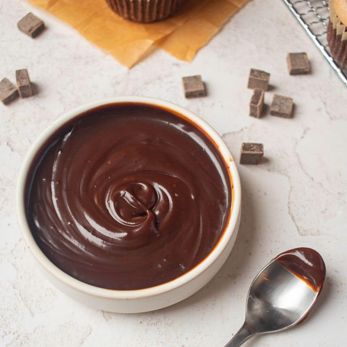 A bowl of smooth chocolate ganache with a spoon beside it, surrounded by chocolate chunks and baked goods in the background.