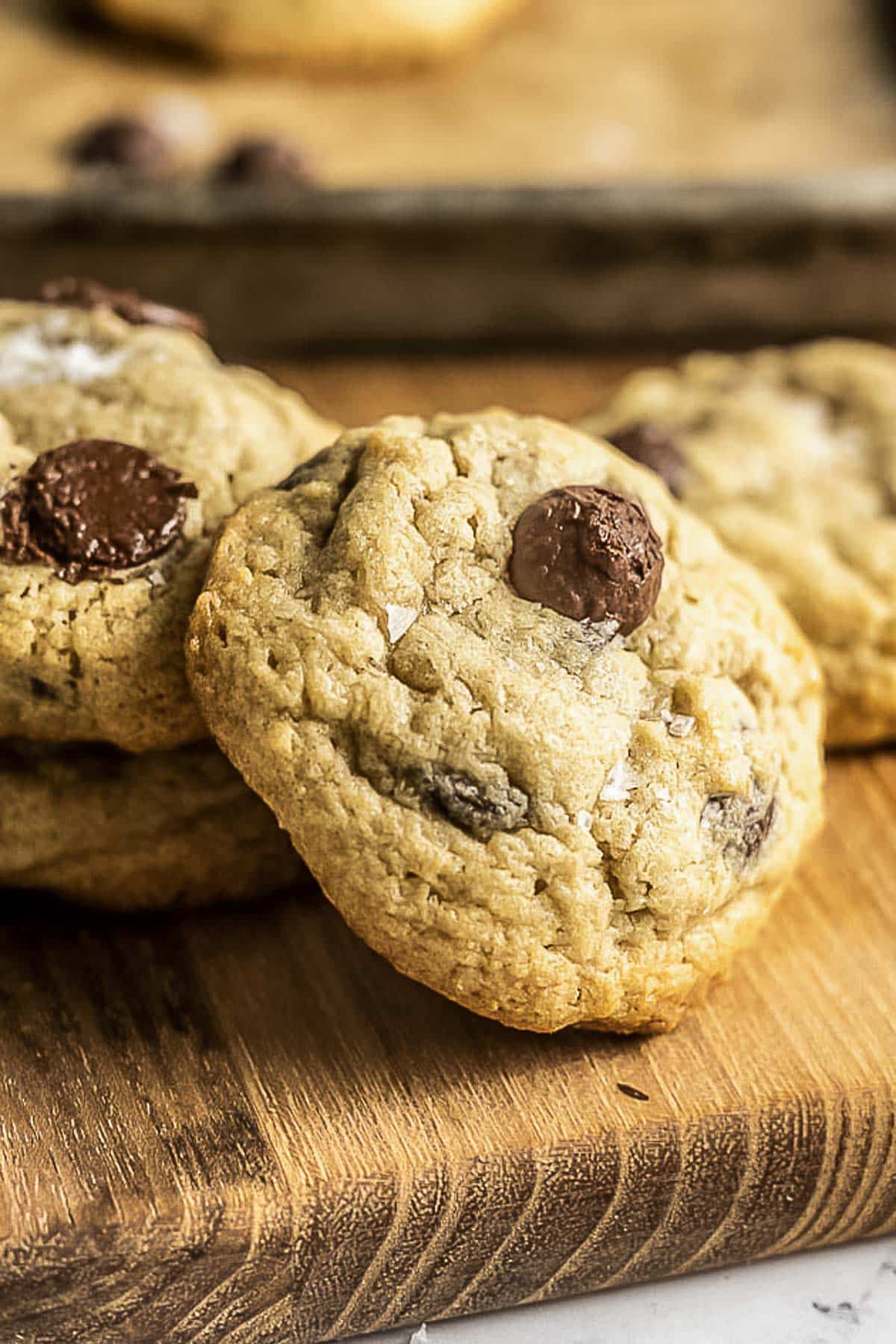 Close-up of thick chocolate chip cookies on a wooden board, with one cookie topped by a single chocolate chip.