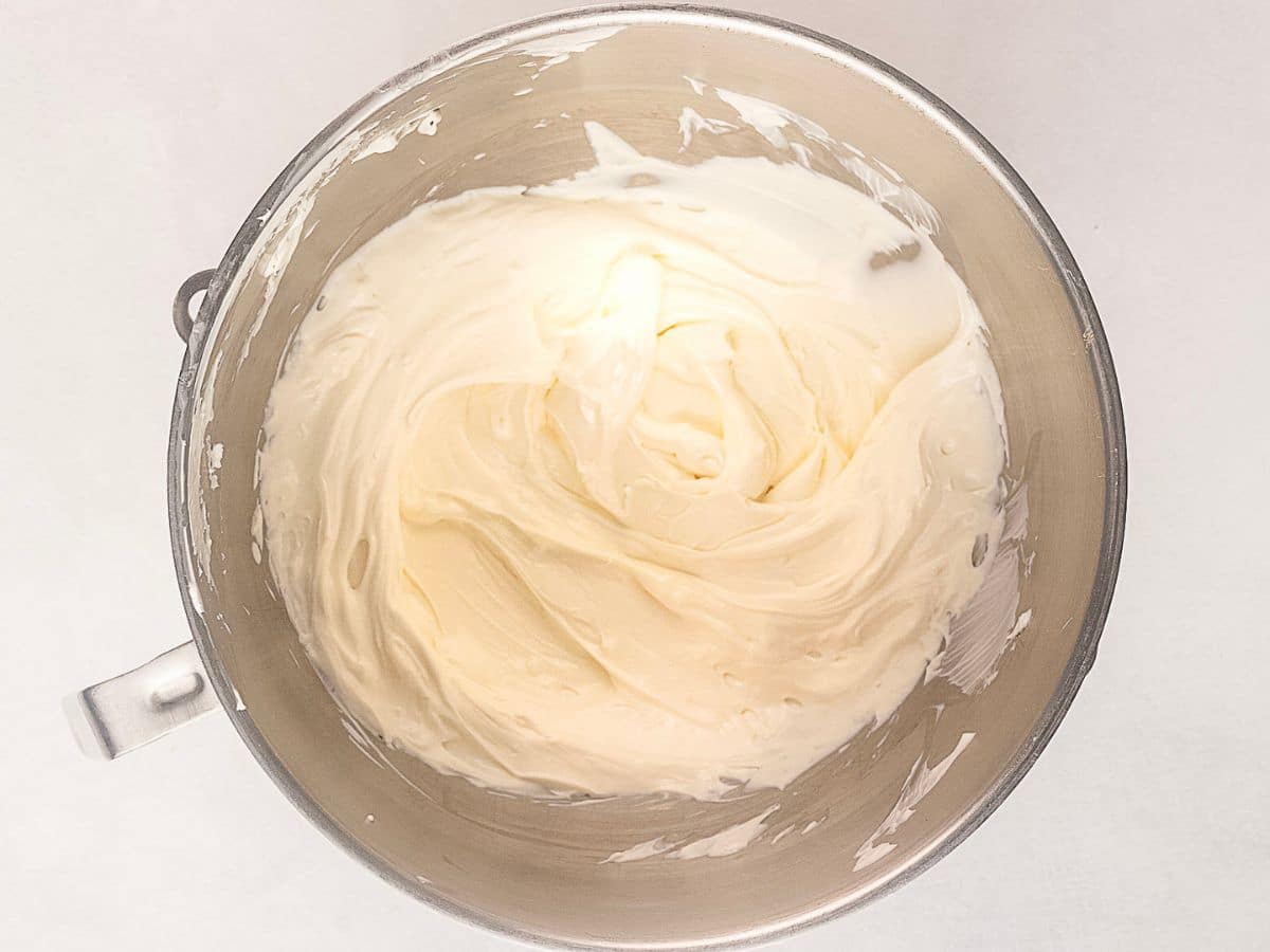 A metal mixing bowl filled with smooth, creamy white frosting against a plain white background.