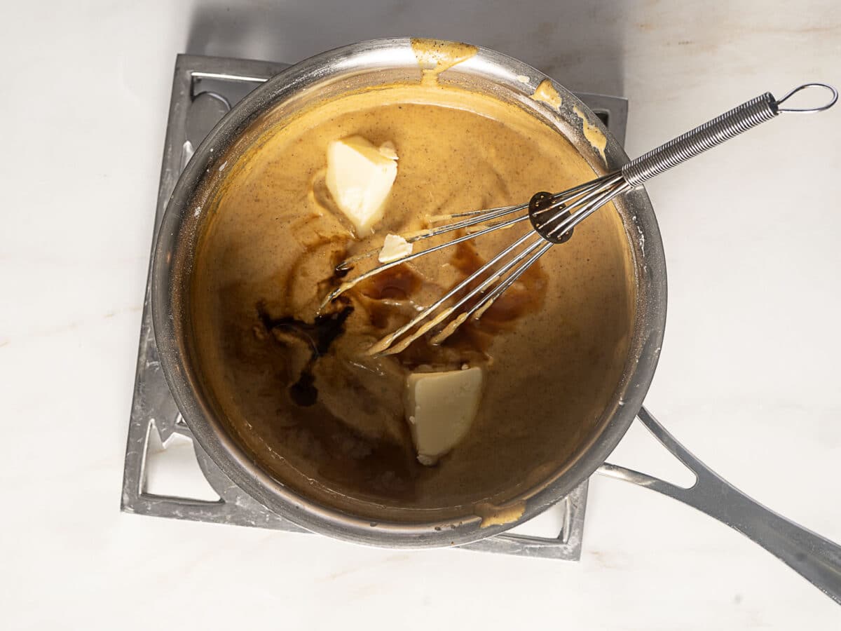 A metal saucepan with a whisk contains a brown sauce mixture, with pieces of butter and a splash of vanilla being mixed in, on a white countertop.