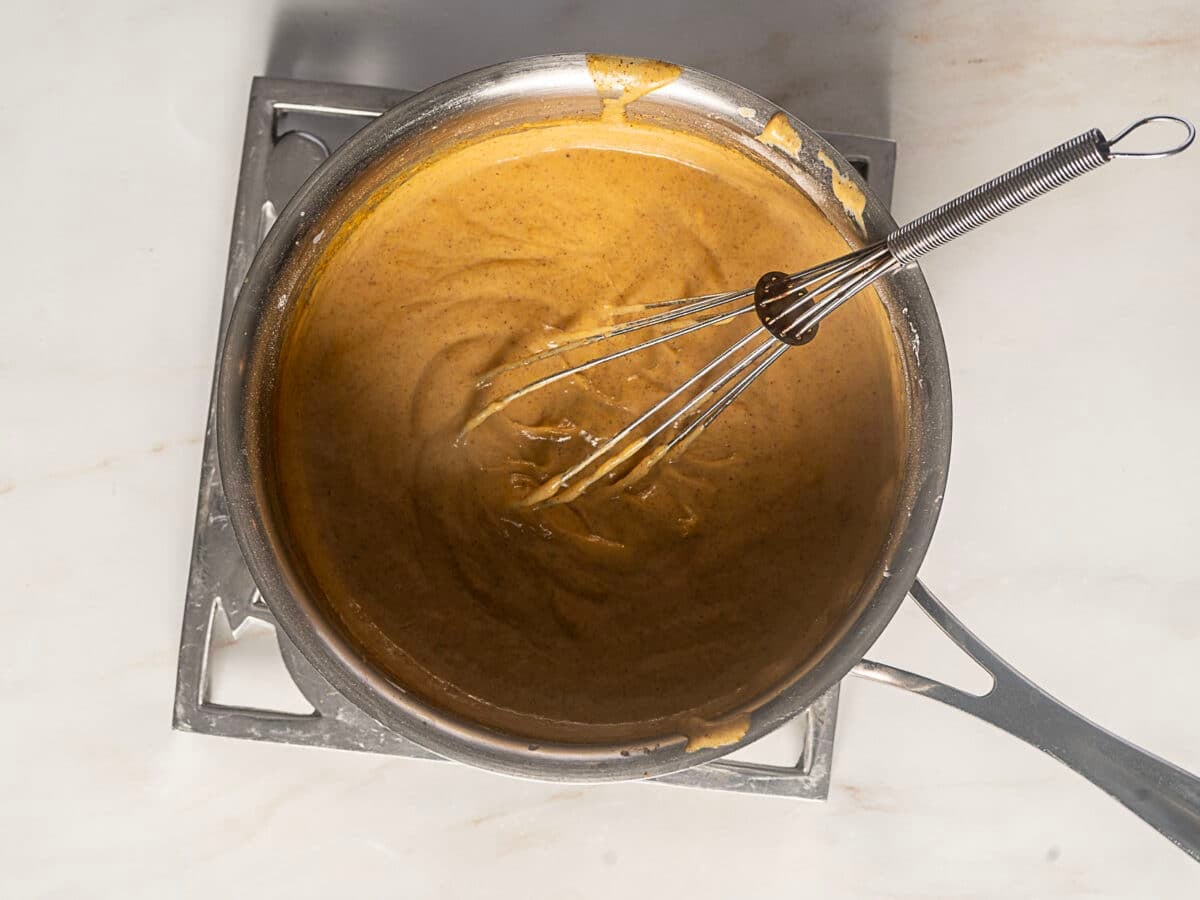 A metal whisk rests in a saucepan filled with thick brown gravy, sitting on a metal trivet on a light-colored countertop.