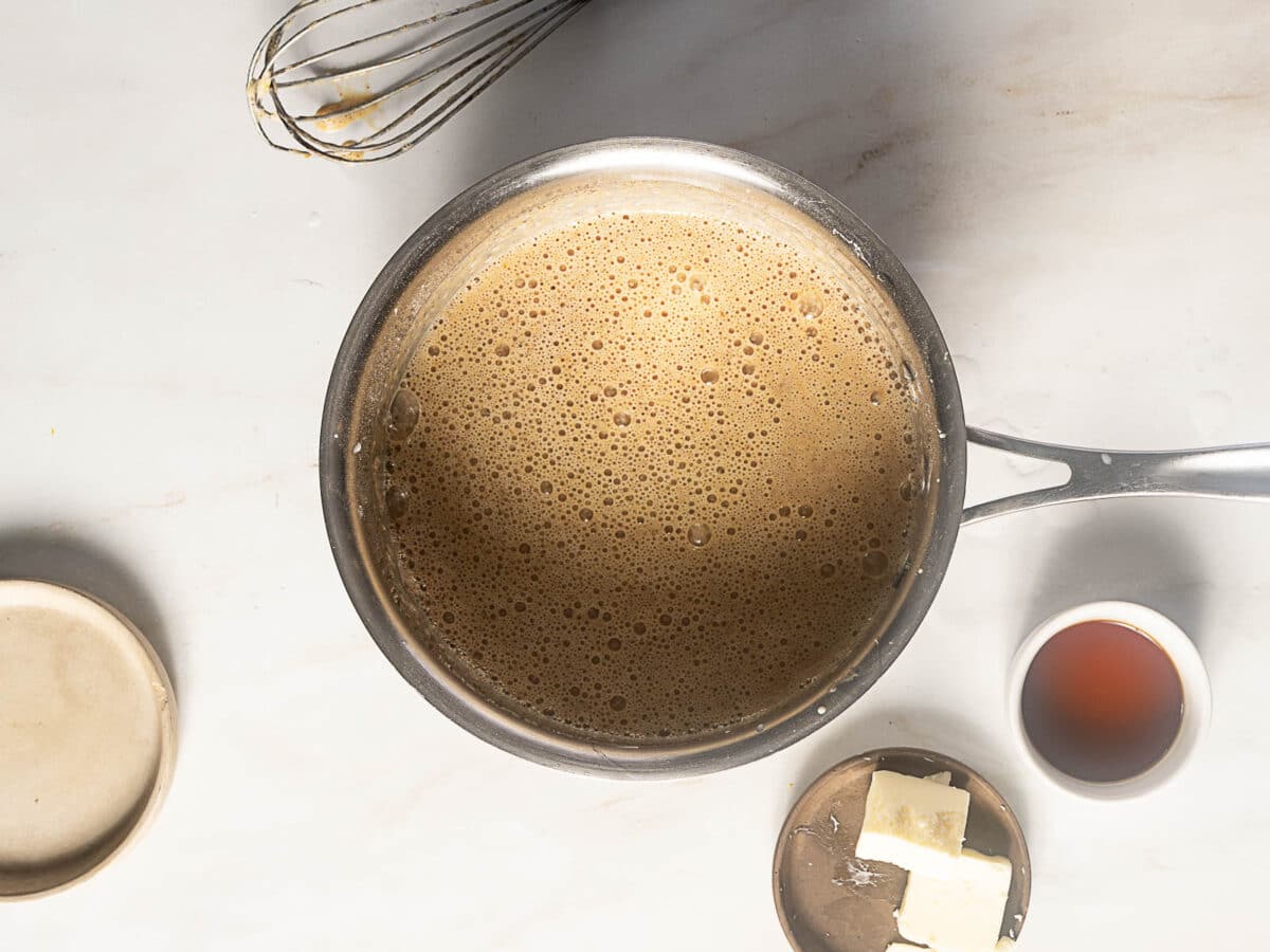 A saucepan filled with a frothy brown mixture sits on a countertop next to a whisk, a small cup of brown liquid, a dish of butter, and a ceramic lid.