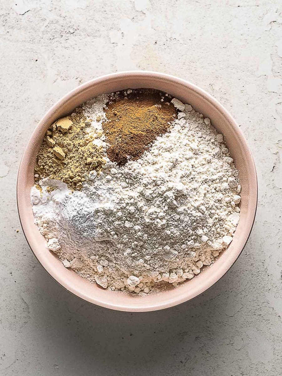 A pink bowl filled with flour, baking powder, baking soda, ground ginger, cinnamon, and other dry baking ingredients on a light countertop.