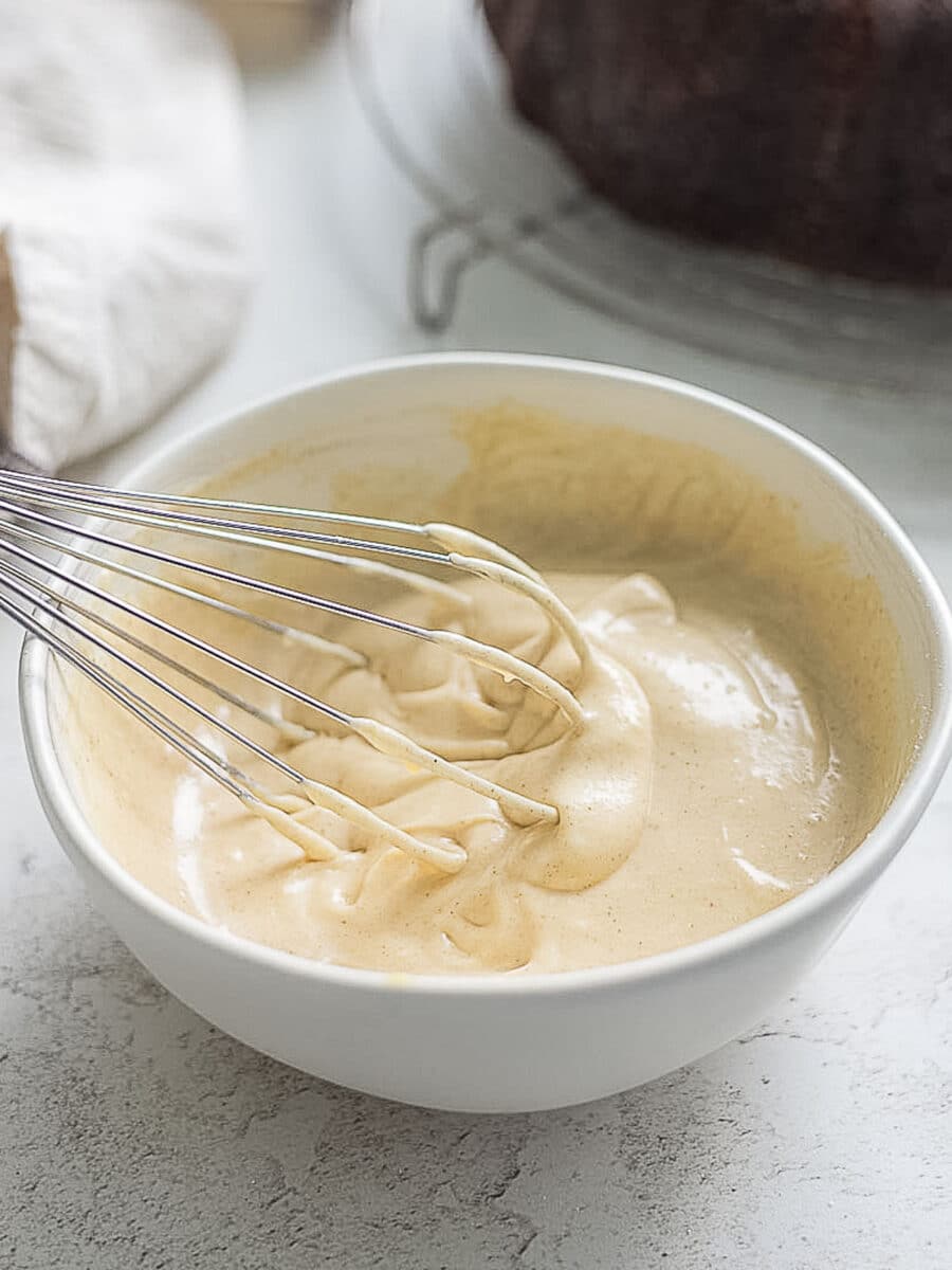 A metal whisk resting in a white bowl filled with creamy, light brown batter on a light-colored countertop.